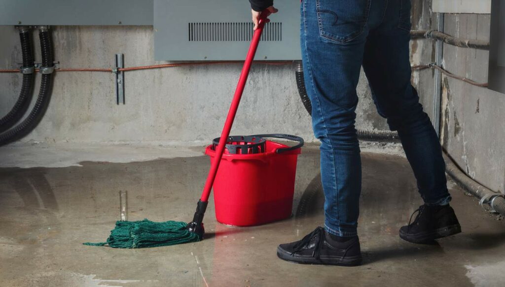 A person in jeans and black shoes mops water from their basement floor